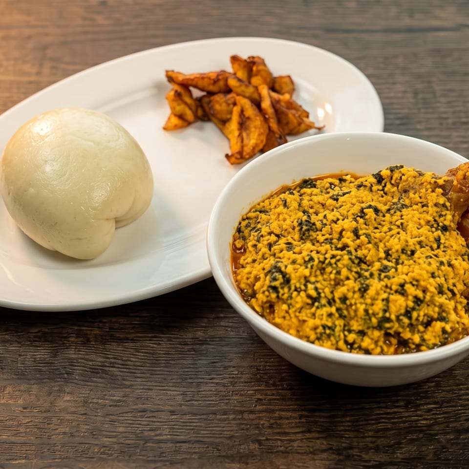 A bowl of egusi soup with chicken, a side of pounded yam, and fried plantains on a wooden table.
