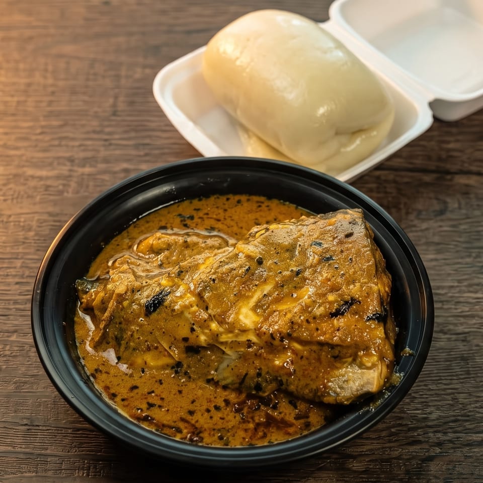 A black bowl of stewed fish in a rich, orange sauce, with fufu in a takeout container.