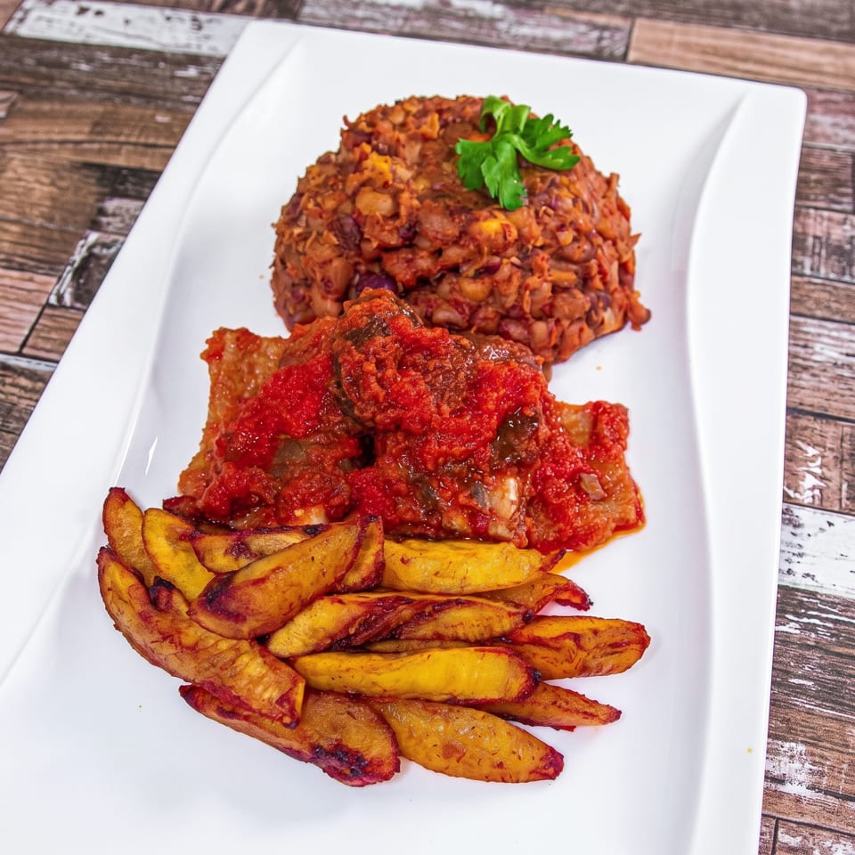 A white rectangular plate holds a meal of red stew, beans, and fried plantains.