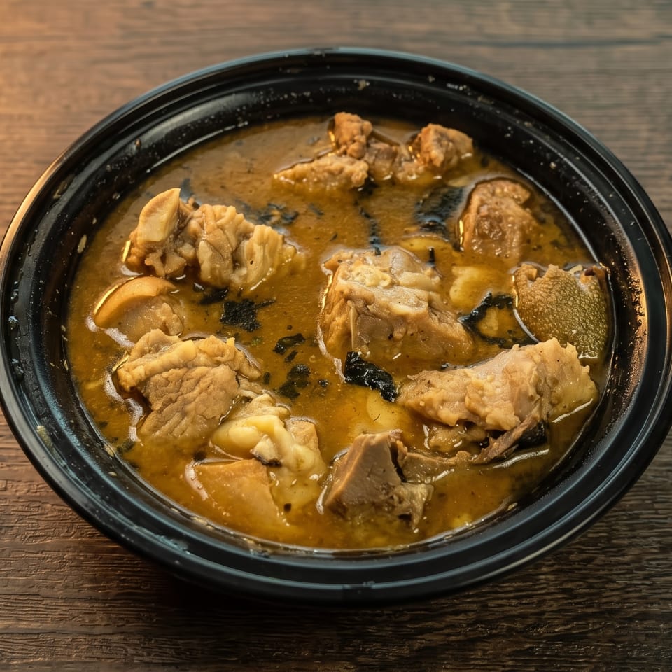 Hearty stew with various meats and tripe in a black bowl on a wooden table.