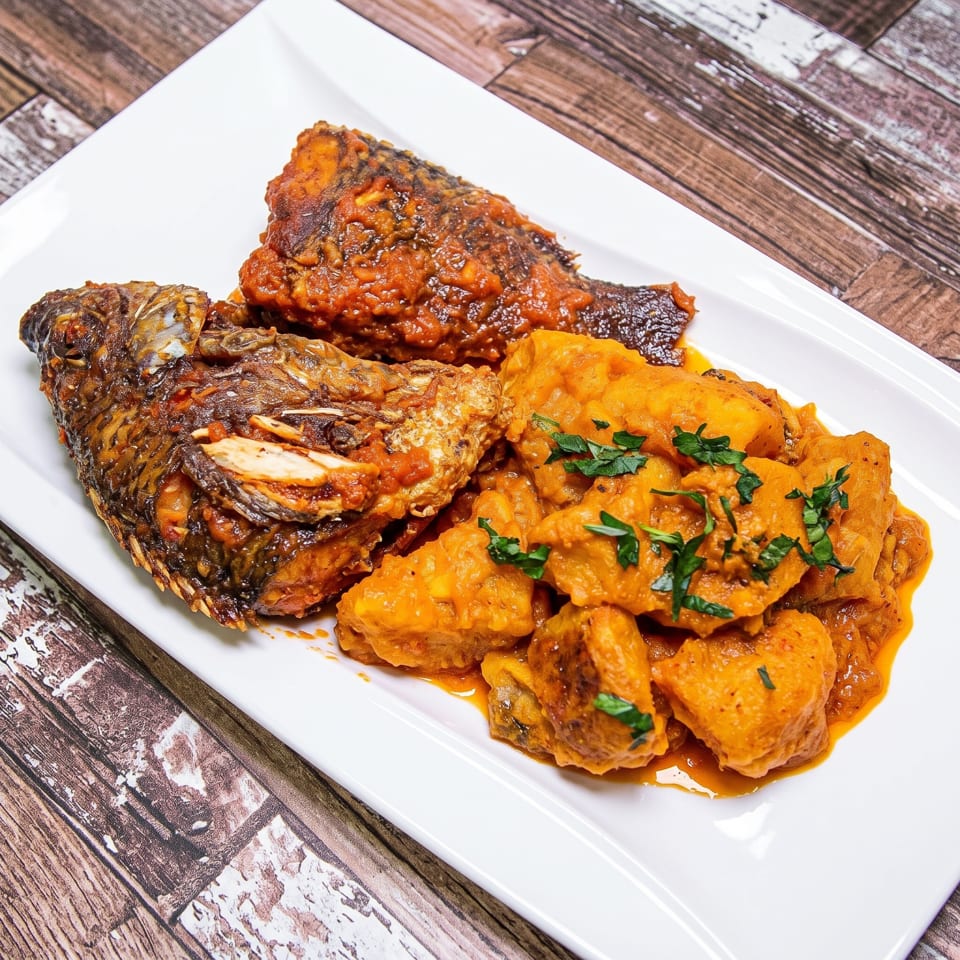 Two whole fried fish covered in red sauce, served with orange stewed yam on a white plate.